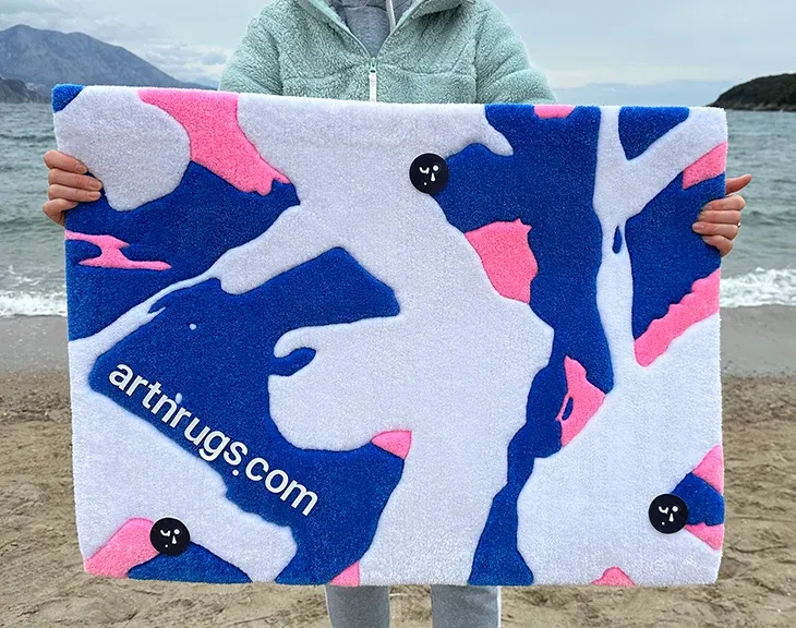 artisan Ksenia Derr holding her handmade blue white pink abstract rug on a beach