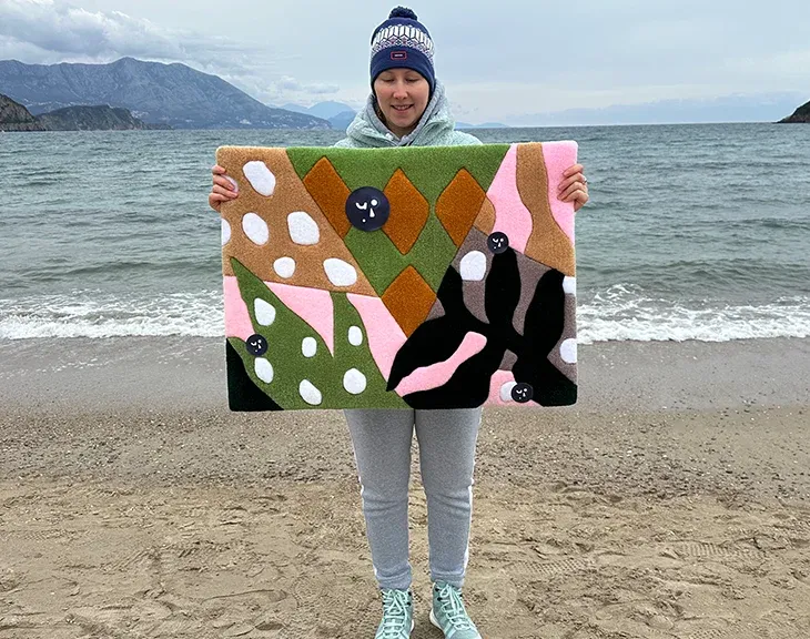 Person holding the olive, pink and mustard leaf rug by the sea.