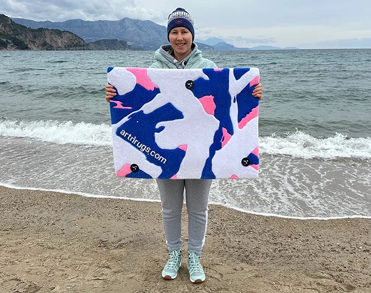 portrait of Ksenia Derr holding her artisan blue white pink abstract rug at shoreline