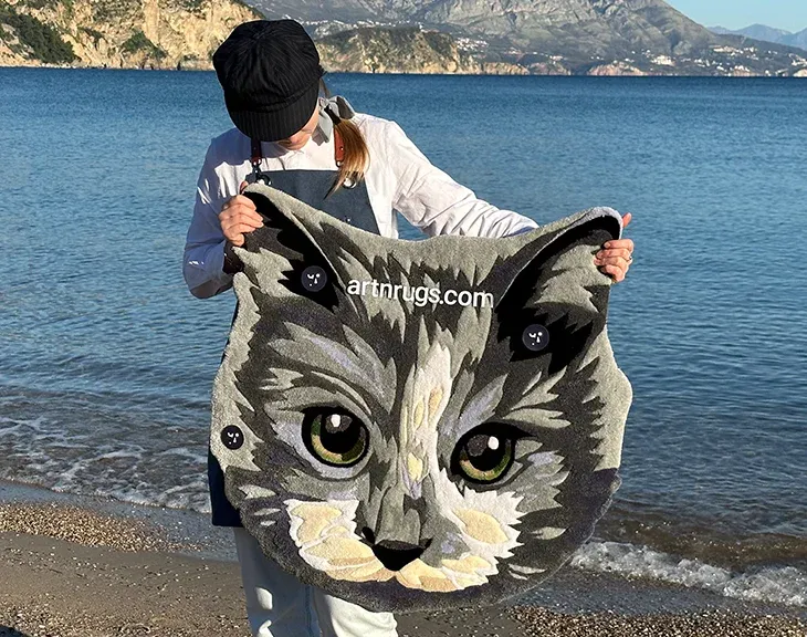 Artesano mostrando alfombra artesanal con retrato de gato gris detallado a la luz del sol junto al mar
