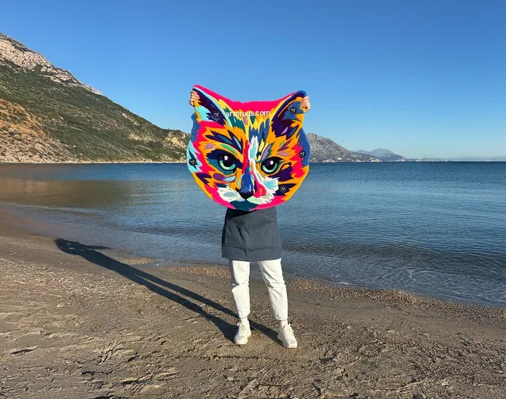 Person holding the multicolor cat portrait rug in sunlight by the sea.