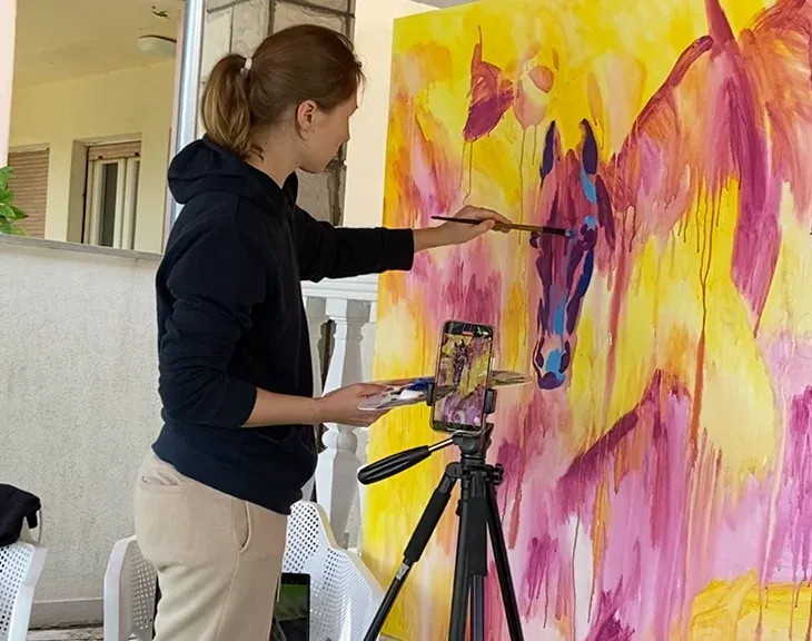 Artist painting a large, colorful horse portrait on canvas in a sunlit studio.